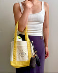 A person wearing a white tank top and purple pants carries a canvas tote bag with yellow handles and a yellow bottom panel. The bag has a front pocket with the text 'SUNNY TIES' and a small black pouch with keys hanging from the side.