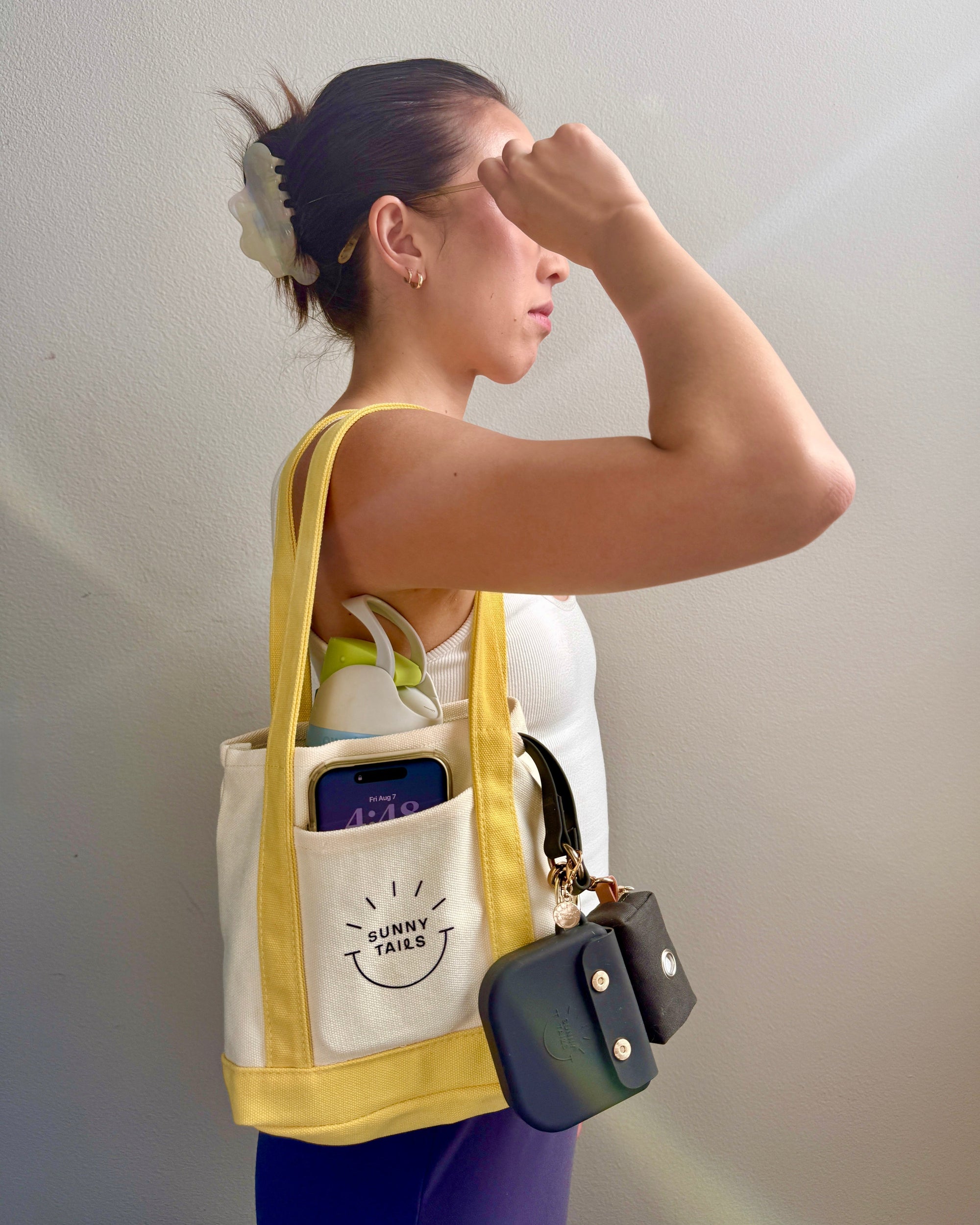 A woman in a white tank top and purple leggings stands in profile, wearing a cream canvas tote bag with yellow trim and a front pocket labeled 'SUNNY SIDE'. The tote bag contains a green bottle and a phone, dog leash, treat pouch, and waste bags.