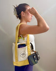 A woman in a white tank top and purple leggings stands in profile, wearing a cream canvas tote bag with yellow trim and a front pocket labeled 'SUNNY SIDE'. The tote bag contains a green bottle and a phone, dog leash, treat pouch, and waste bags.